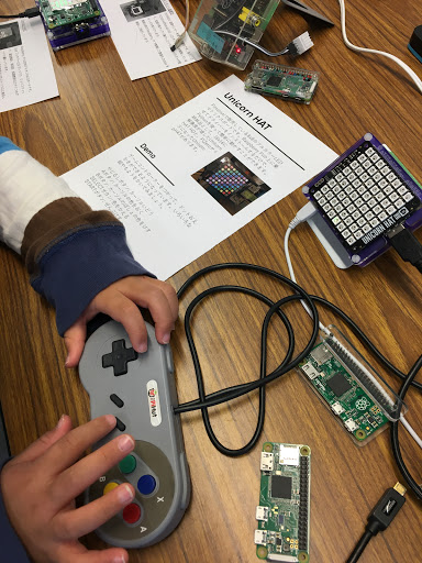 Raspberry Pi ユーザーグループのブースで、子供がゲームパッドを握りラズパイ基板やUnicorn HATのLEDマトリクスに触れる様子